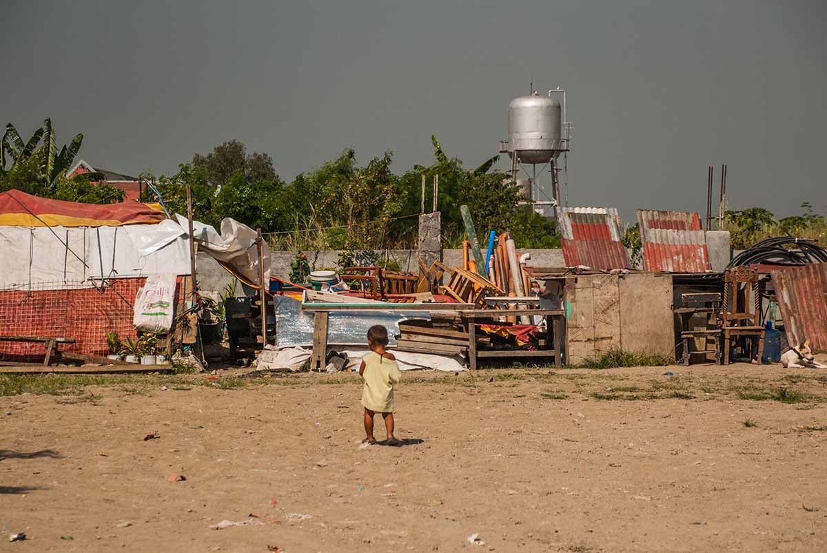 A child stands on an open dirt area with a refugee camp in the background
